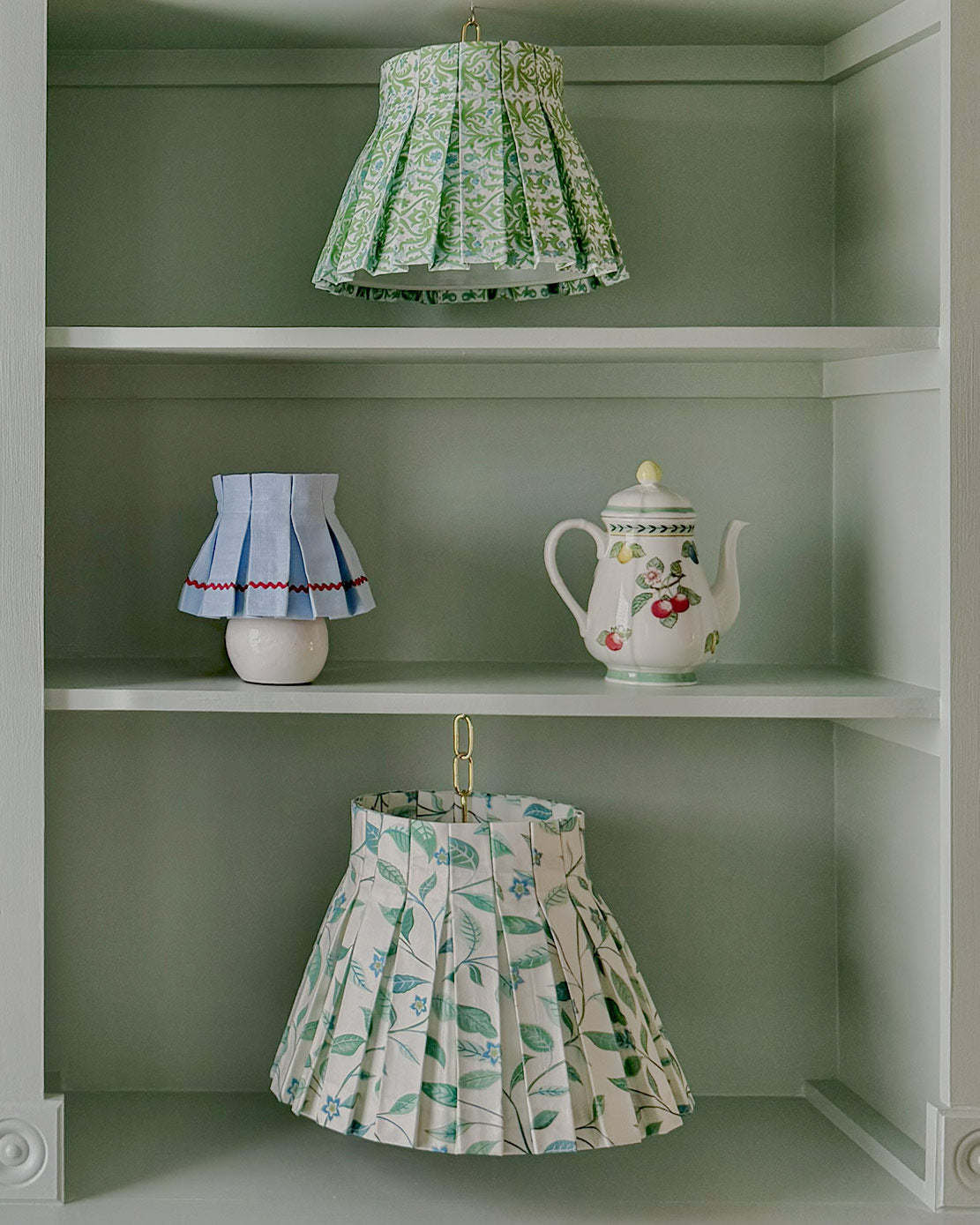 Decorative lampshade skirt in floral prints and a teapot on a shelf against a green wall. Elysse and Siena.