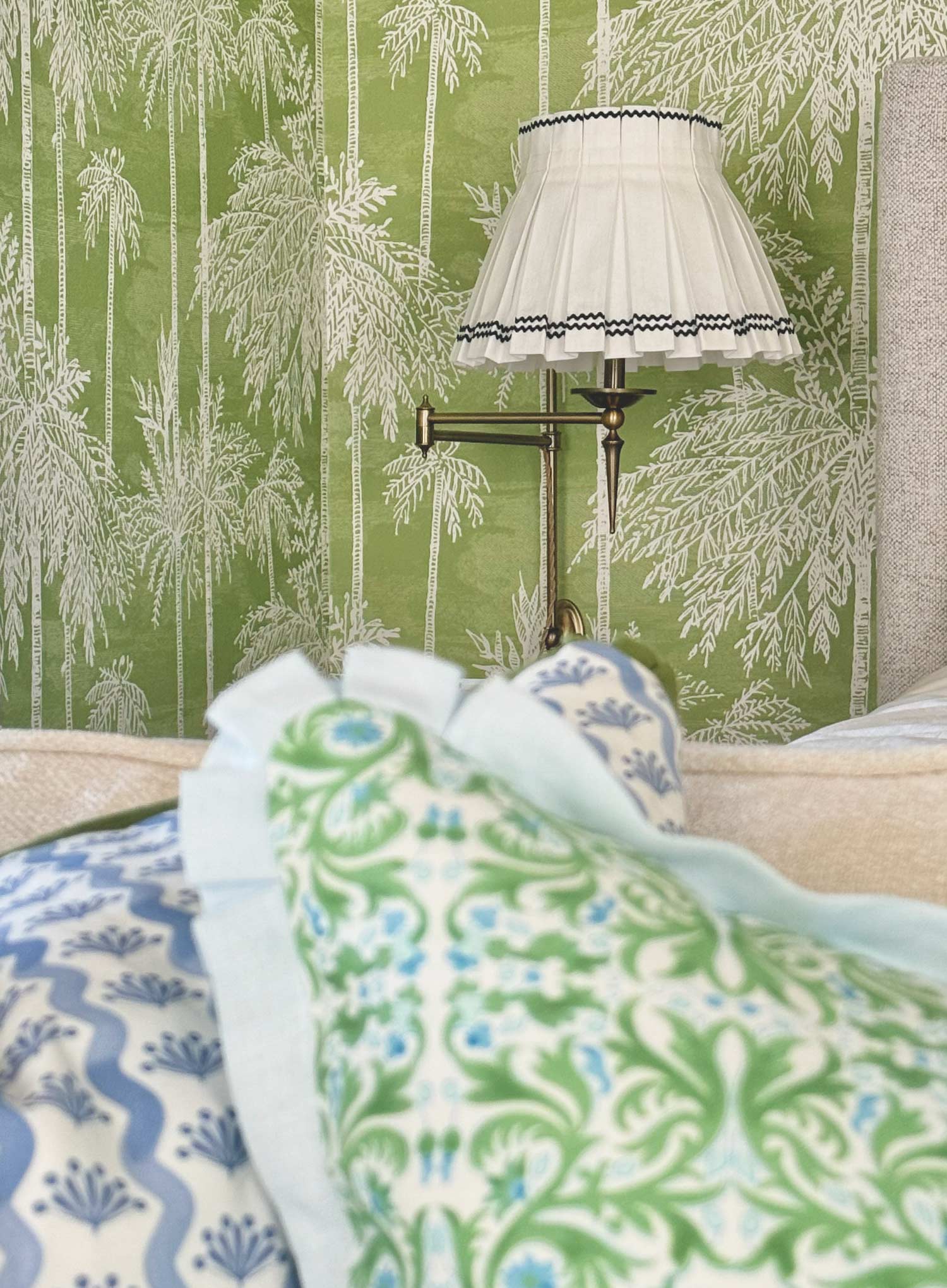 A styled bedroom corner featuring green and white patterned wallpaper, a brass wall lamp with a box pleated white shade with navy ric rac trim, and a bed in the foreground with blue and green patterned siena designer pillows.