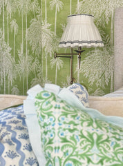 A styled bedroom corner featuring green and white patterned wallpaper, a brass wall lamp with a box pleated white shade with navy ric rac trim, and a bed in the foreground with blue and green patterned siena designer pillows.